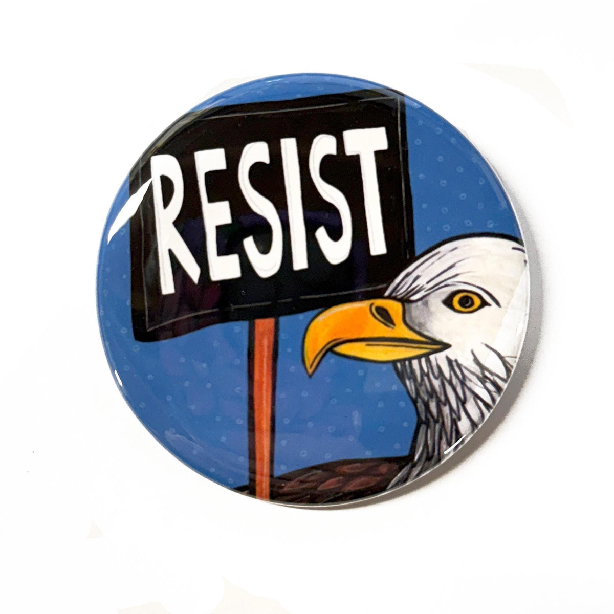 round pin or magnet with a bald eagle holding a resist protest sign on a blue background