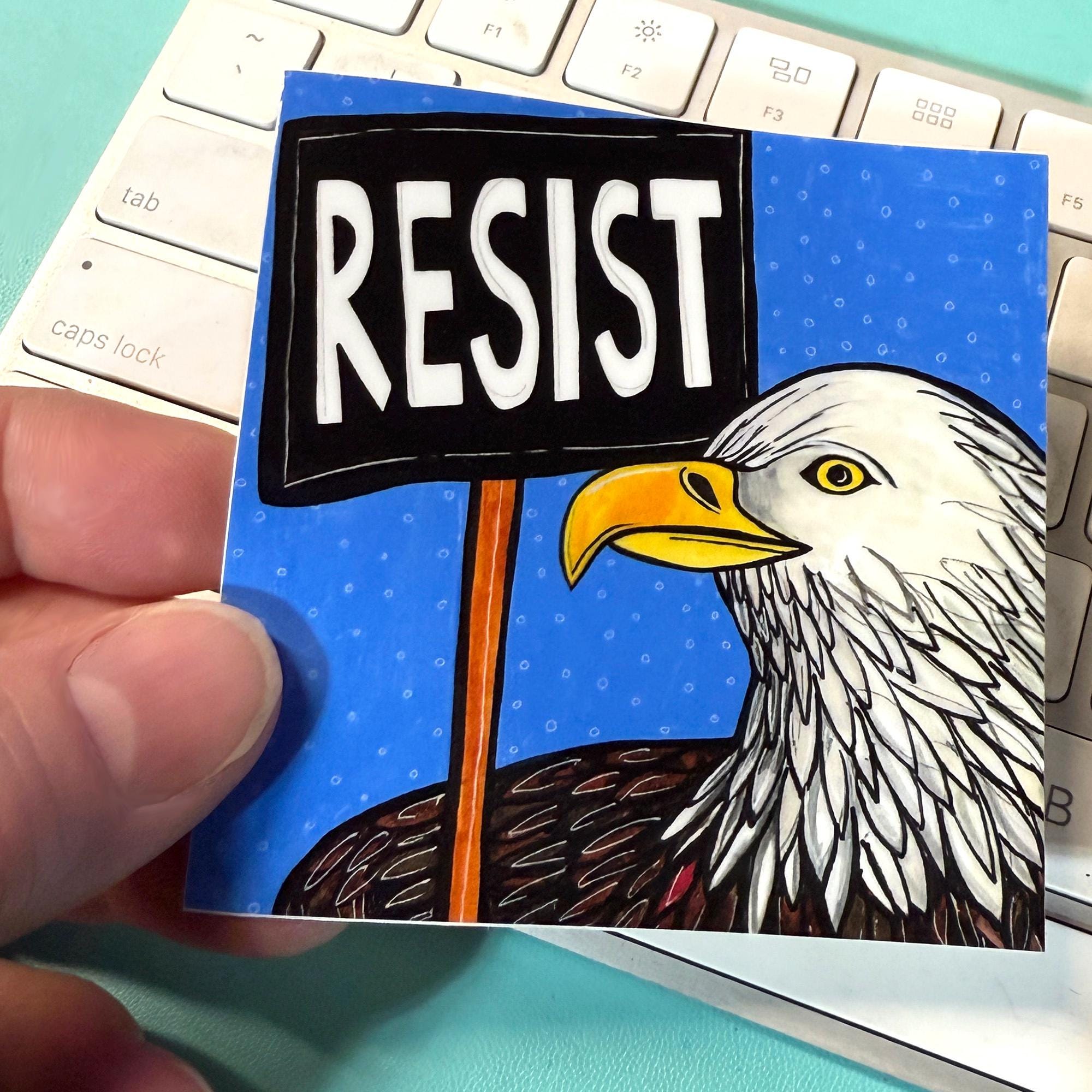 Hand holding Bald Eagle Resist sticker. Sticker is square with hand drawn bald eagle on a blue background holding a RESIST protest sign.