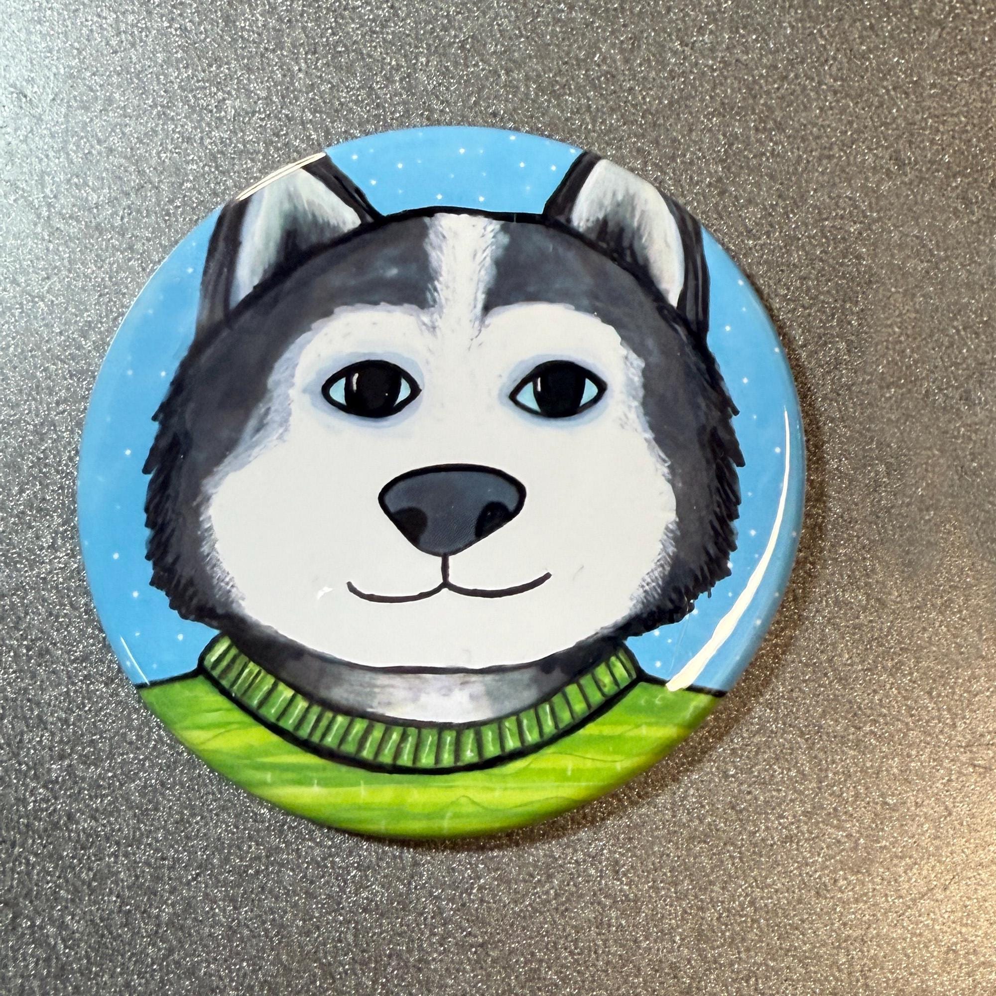 Round siberian husky pin or magnet features gray and white husky with blue eyes wearing a bright green striped sweater with a light blue background with white snow flurries. Shown on a gray metal surface.