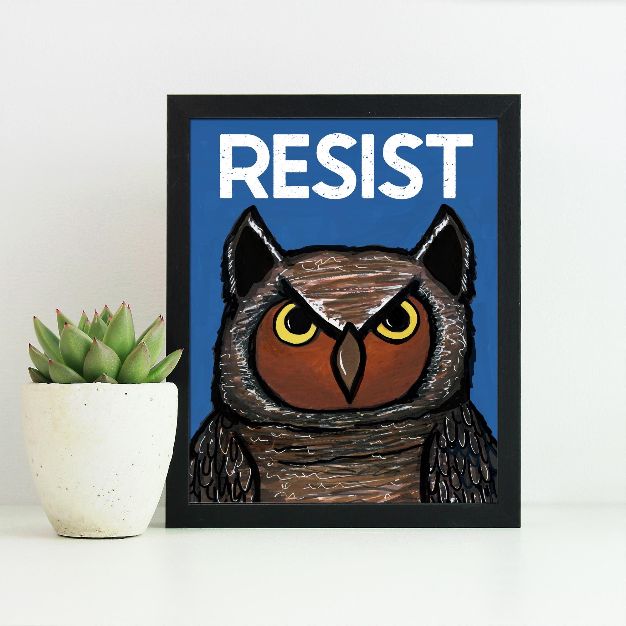 Wise Owl Resist art print in a black frame sitting next to a succulent plant in a white pot. The art features a hand drawn owl on a blue background with the words resist in white above its head.