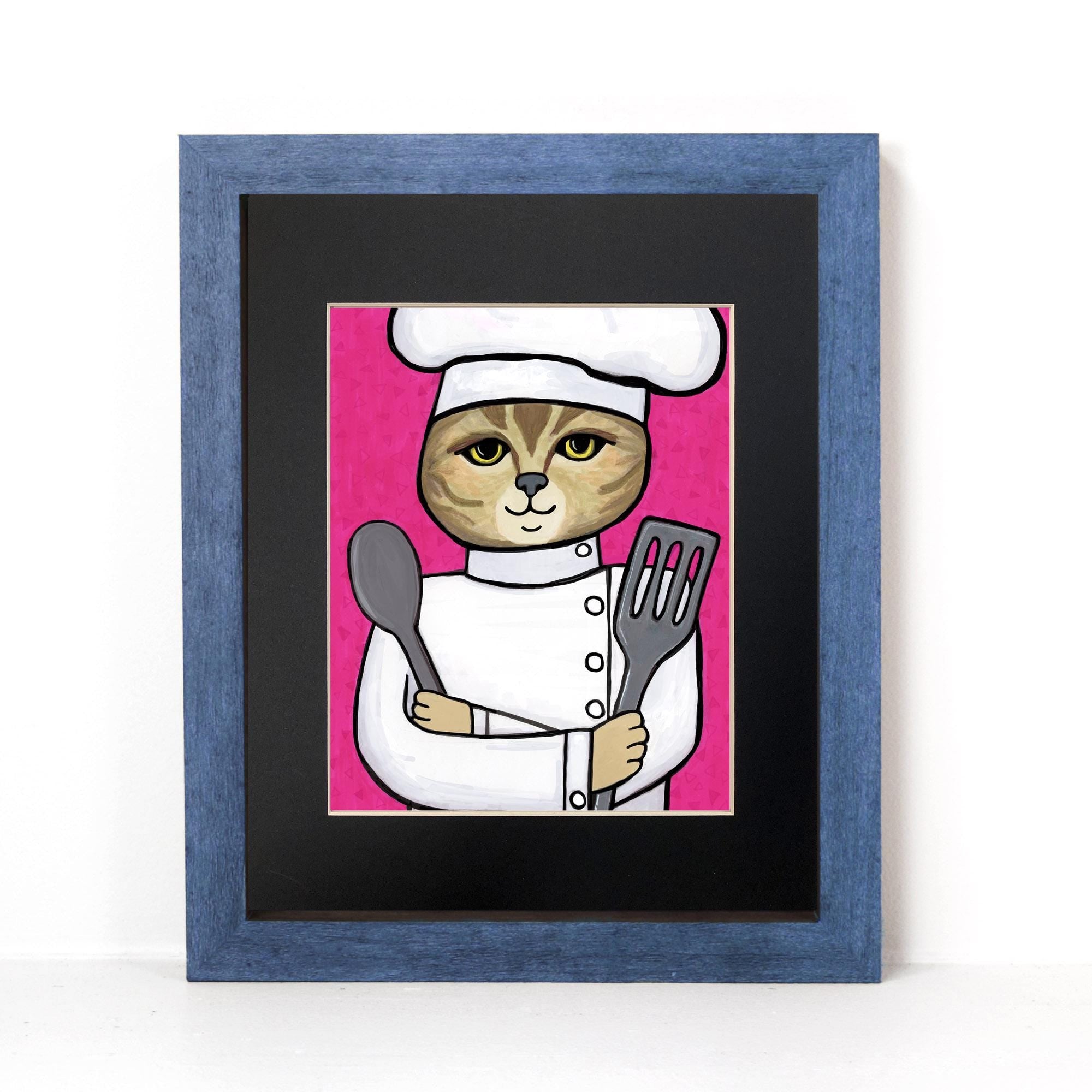 Cat Chef art print in a black mat and blue wood frame. Art features an american shorthair cat in a chef's hat and jacket holding metal spoon and spatula on a pink background