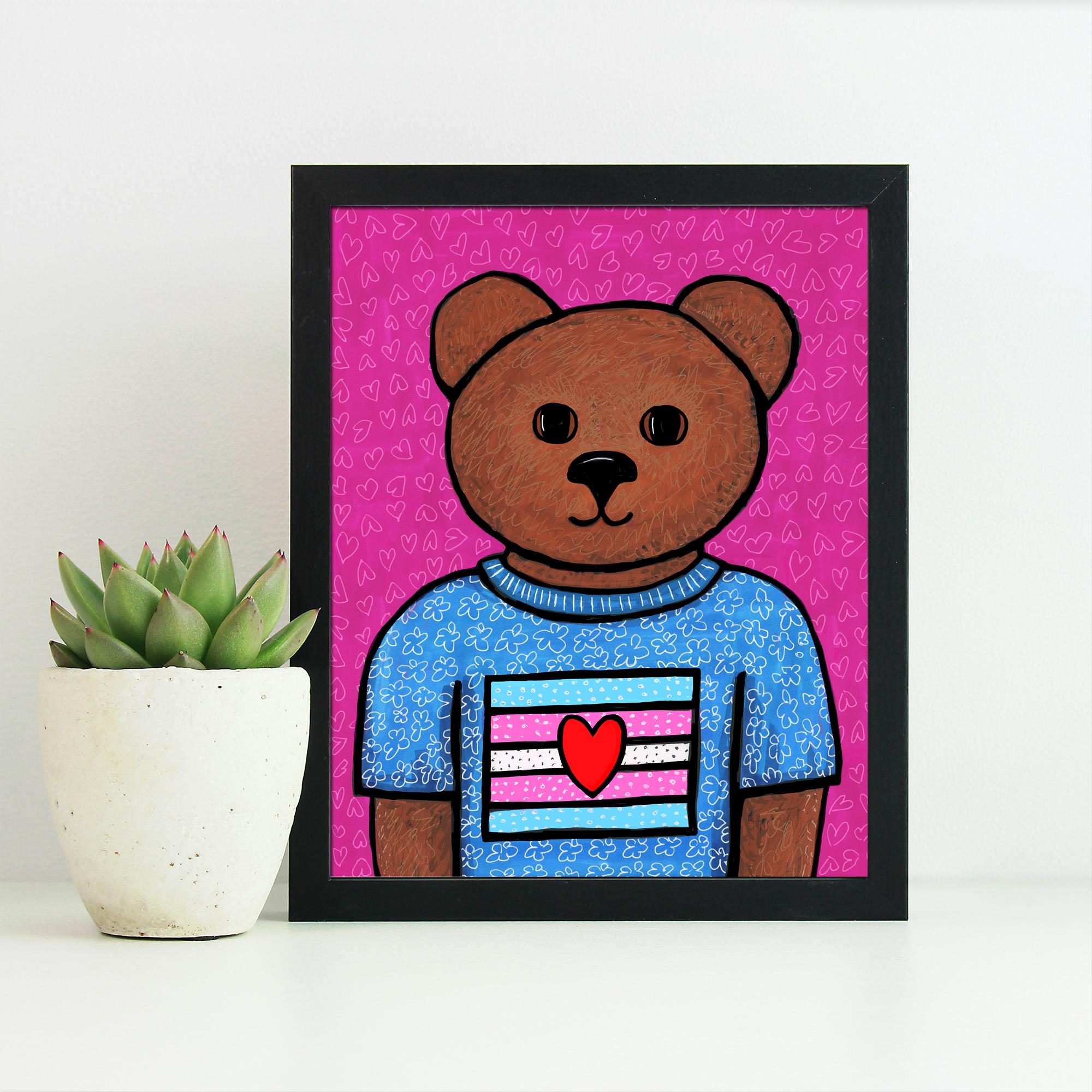 Framed art print in a black frame with potted succulent plant.  It features Rowan - a brown in a blue floral t-shirt with trans flag with a cute heart. Rowan stands against a pink background with little hearts.