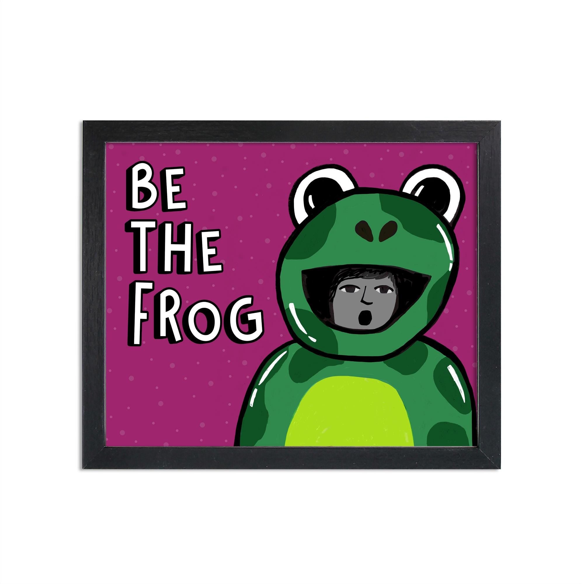 Be The Frog print in a black frame. The design features a deep magenta background with a hand drawn frog inflatable costume with a person inside it and the words Be The Frog in a white font with black shadow.