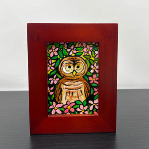 Black table with a small framed acrylic painting of a brown barred owl surrounded by pink apple blossoms and green leaves