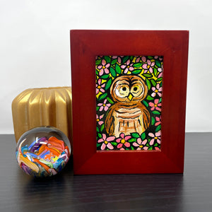 Barred Owl painting in cherry wood frame on black table next to paperweight and candle holder. Acrylic painting of a brown barred owl surrounded by pink apple blossoms and green leaves