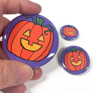 Hand holding 2.25 inch pumpkin pin next to 1.25 inch pin and 1 inch pin