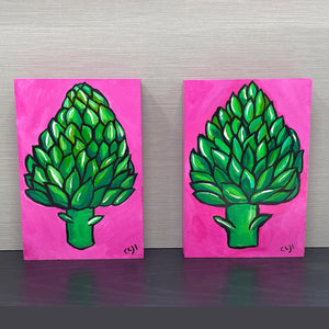 pair of artichoke paintings on black shelf with gray background. paintings feature green artichoke on pink background.