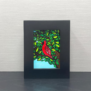 Small cardinal painting in a black frame sitting on a black shelf with gray background