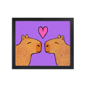 Capybaras in Love art print set in black frame (framed not included). Two brown capybaras with eyes closed facing each other with pink heart above them. Background is purple with small dots.