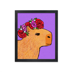 Capybara in Rose Crown art print shown in black frame. Art features a side view of a brown capybara with eyelashes with a rose crown with red and pink petals and green leaves.