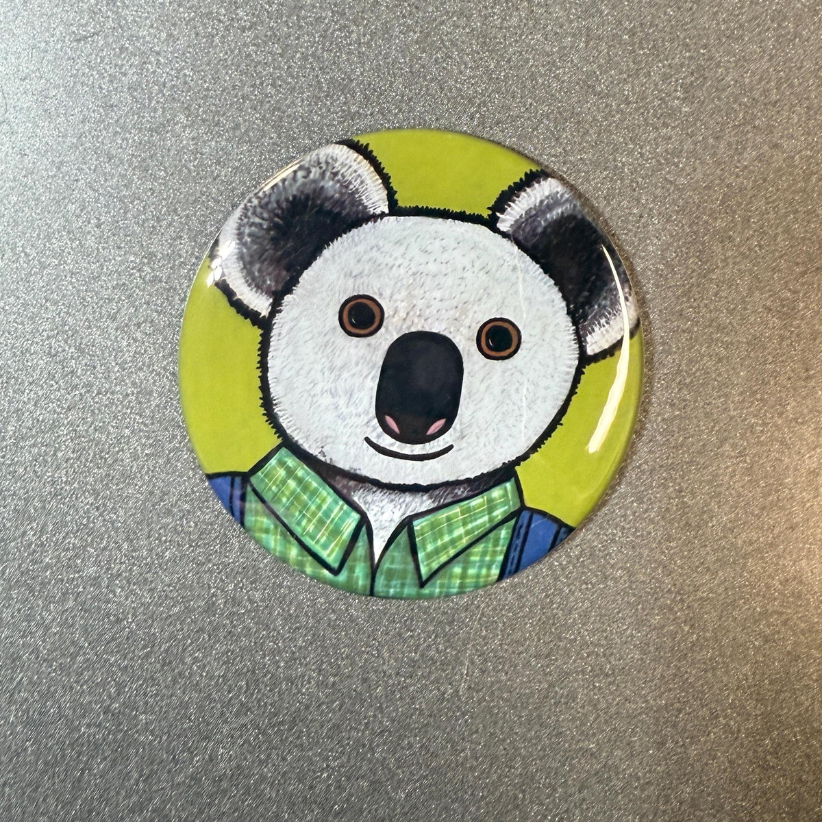 Round button design featuring koala in green plaid shirt and blue overalls on a chartreuse background. Available as pins, magnets, and pocket mirrors.