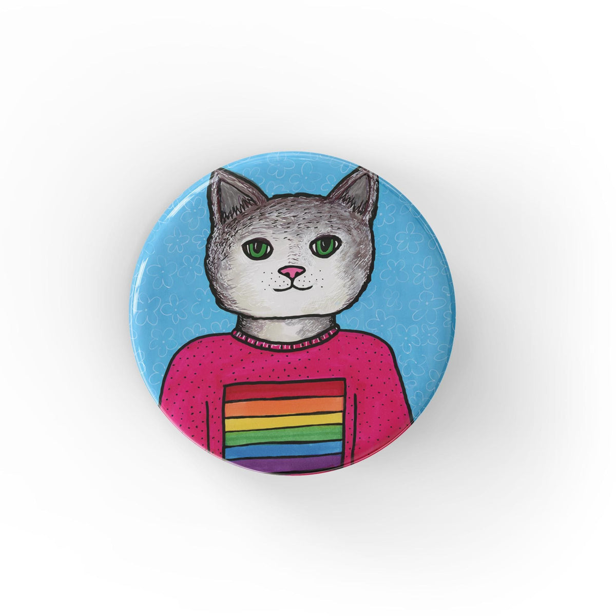 Round pin back button or fridge magnet featuring a gray and white cat in a pink shirt with a rainbow flag on it. The background is blue with white floral design.
