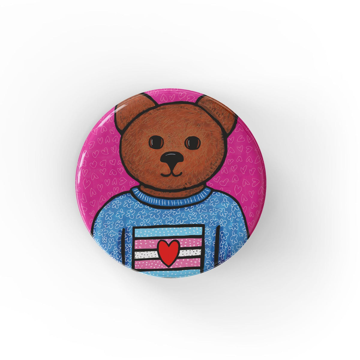 Round pink button featuring a brown bear in a blue floral shirt with trans flag with a red heart. Available as magnet or pin.