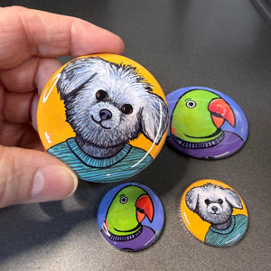 a hand holding the 2.25 inch Millie the Shih Tzu button above other buttons including the 1.25 inch Millie and 2.25 and 1.25 inch Oscar the Alexandrine Parrot - background is metal gray