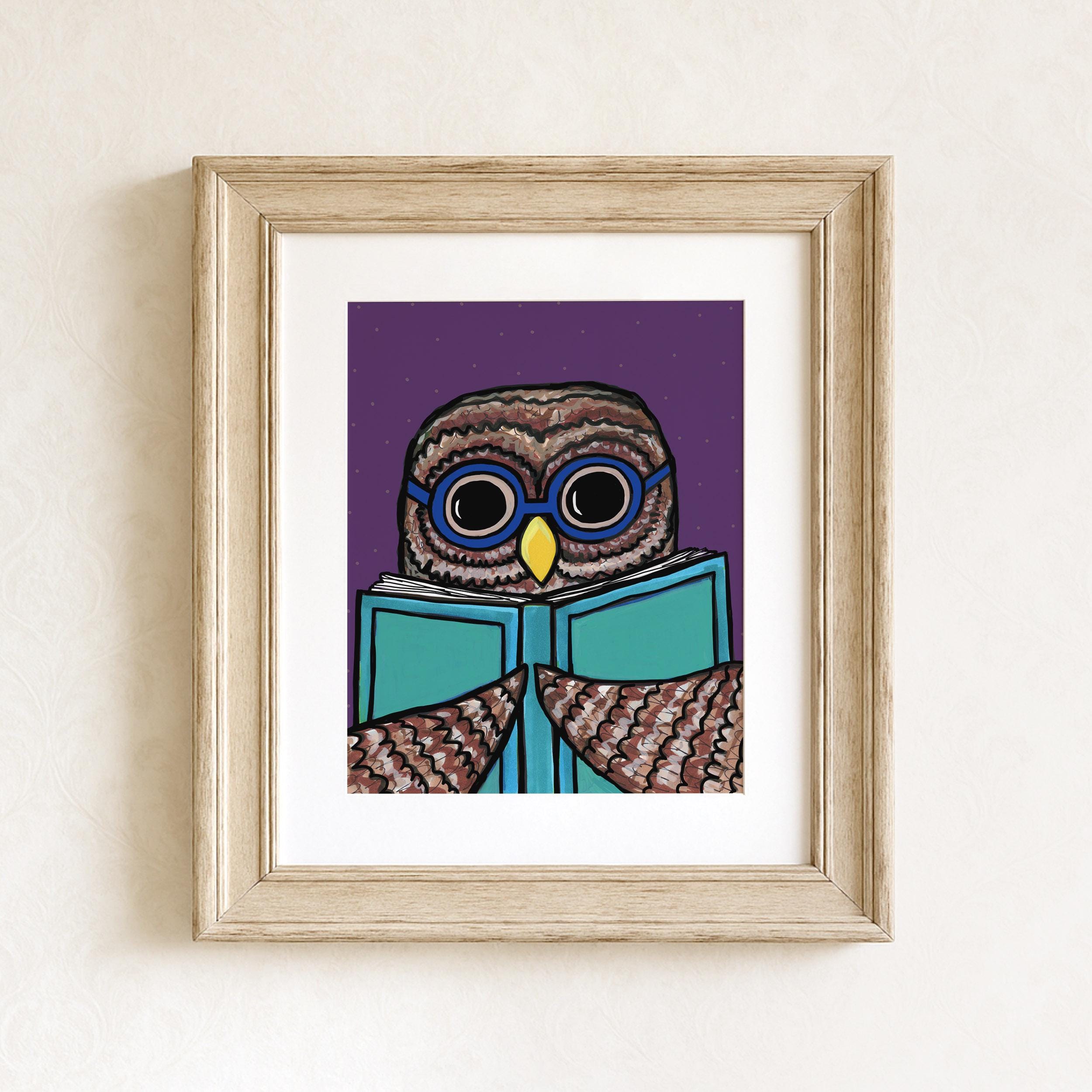 Simon the Owl Reads art print is shown in a wood frame with white matte on a white wall. The owl is wearing blue glasses and holding a light blue book. The background is purple.