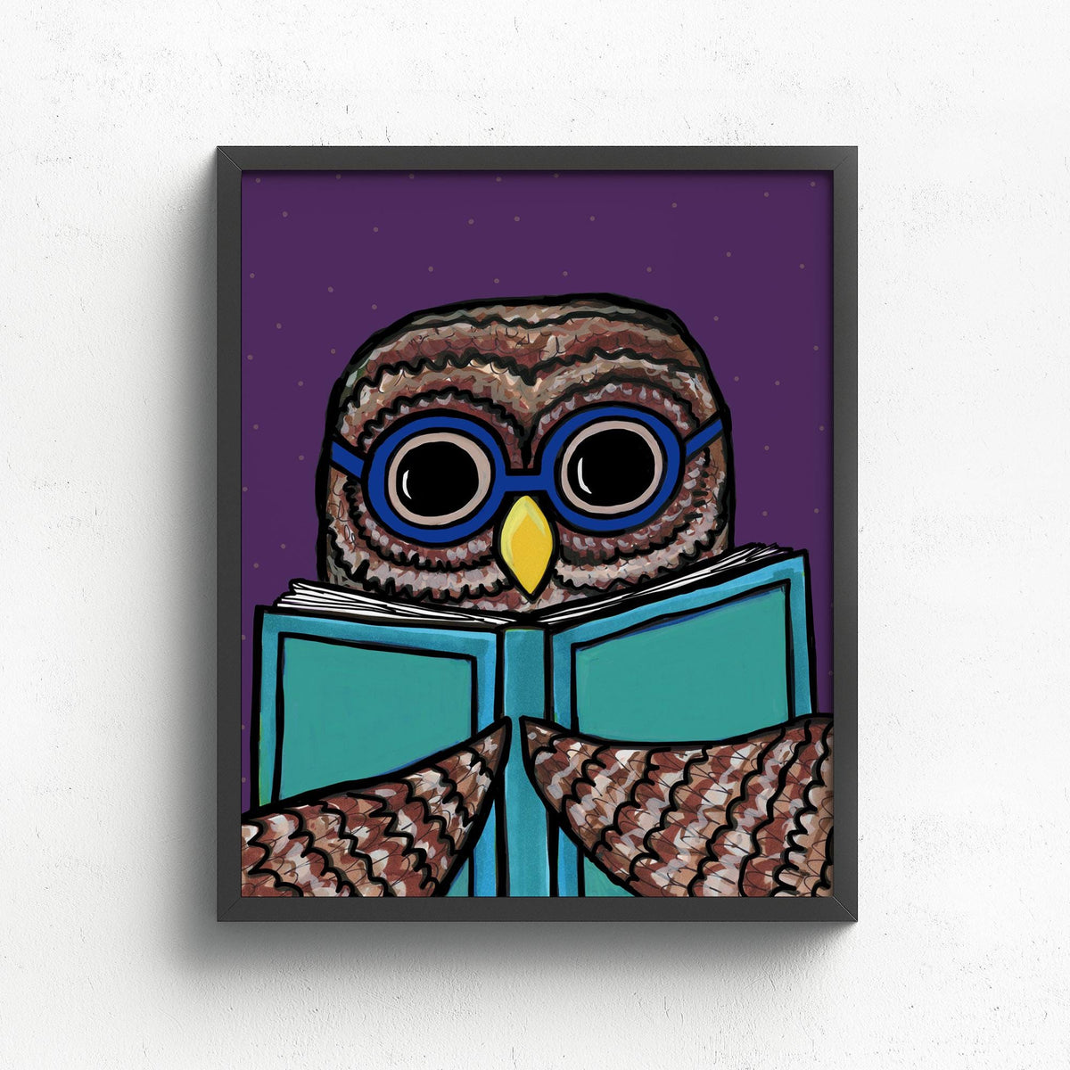 Framed owl art print in a black frame on a white wall. The owl is wearing blue glasses and holding a light blue book. The background is purple.