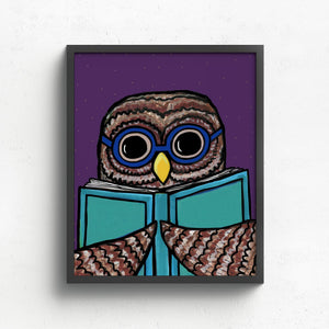 Framed owl art print in a black frame on a white wall. The owl is wearing blue glasses and holding a light blue book. The background is purple.