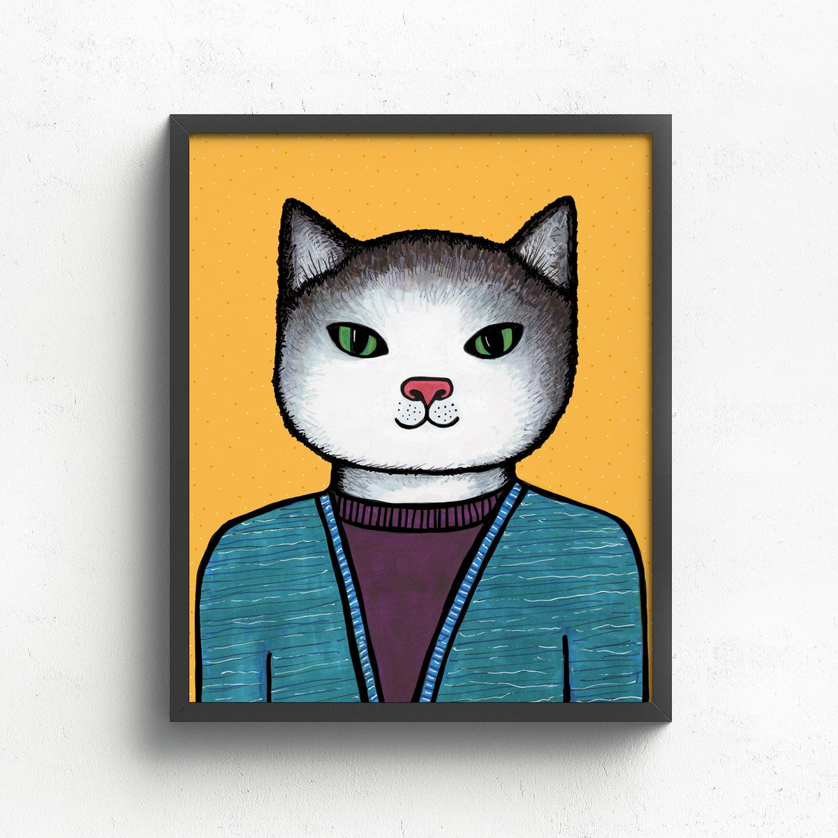 Cute cat drawing print in a black frame on white wall. The cat is wearing a purple shirt and blue sweater and against a yellow background.