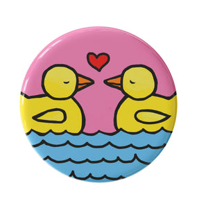 round pink button with two yellow ducks in love. They are facing each other as they sit on blue water. A red heart appears between them above their head level.