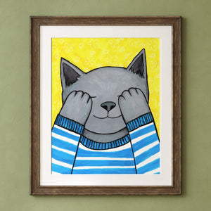 Teddy Plays Hide N Seek art print displayed in a wood frame and white mat shown on a olive green wall. The art shows a gray cat covering eyes with paws. cat is in a blue and white striped sweater. the background is bright yellow with white flower pattern.