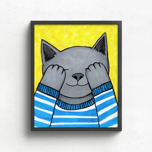 Teddy Plays Hide N Seek art print displayed in a simple black frame on a white wall. The art shows a gray cat covering eyes with paws. cat is in a blue and white striped sweater. the background is bright yellow with white flower pattern.
