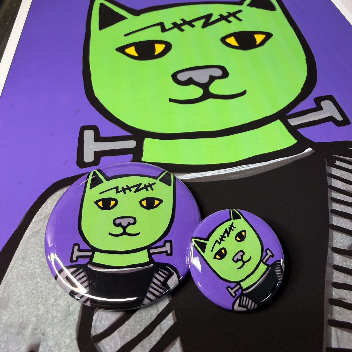 Frankencat buttons shown on larger print of the design. Green cat with forehead scar and neck bolts in a black suit. Background is purple.