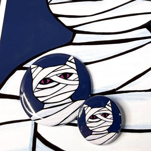 Cat Mummy buttons shown on a Cat Mummy print. Cat Mummy is wrapped in white bandages and has purple eyes. The background is dark blue.