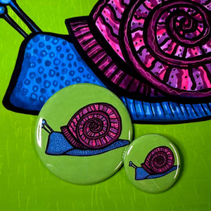 two different size snail buttons - 1.25 and 2.25 inch. Snail is blue with purple spiral shell. Background of buttons are green. Shown on a detail of snail art print.