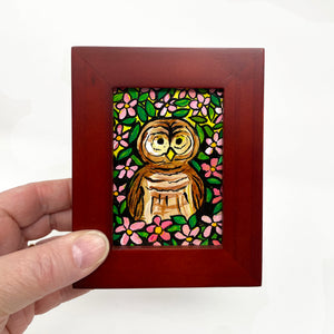 Hand holding a small framed acrylic painting of a brown barred owl surrounded by pink apple blossoms and green leaves