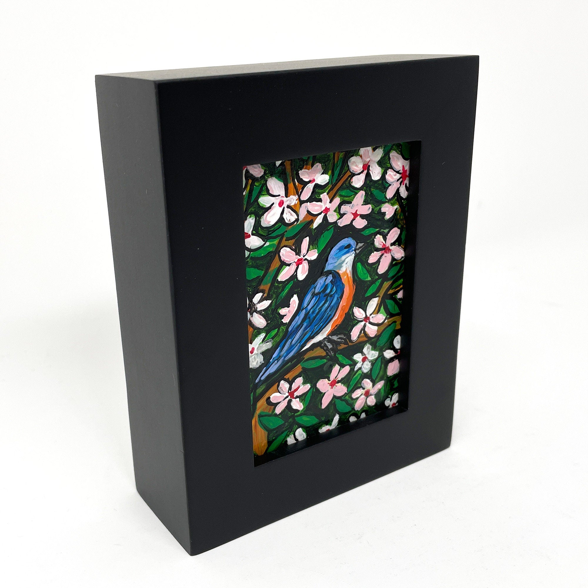 Side view of bluebird painting in black frame. In the painting a bluebird sits on a cherry tree surrounded by pink and white cherry blossoms and green leaves.