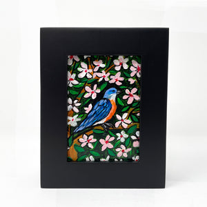 Front view of bluebird painting in black frame. In the painting a bluebird sits on a cherry tree surrounded by pink and white cherry blossoms and green leaves.