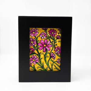 Front view of black wood frame holding pink cosmos flower painting. Flowers and green leaves are on a yellow ochre background.