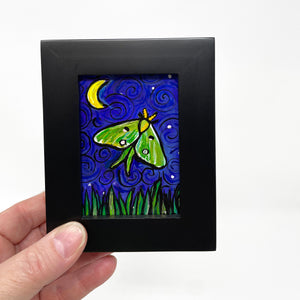 Hand holding Luna Moth painting in black wood frame that features green and yellow luna moth on night sky with crescent yellow moon and white stars. Long grasses dance along the bottom of the scene.