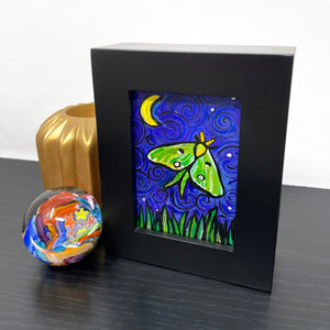 Side view of Luna Moth painting on a shelf with a trinket box and paperweight. Painting in black wood frame features green and yellow luna moth on night sky with crescent yellow moon and white stars. Long grasses dance along the bottom of the scene.