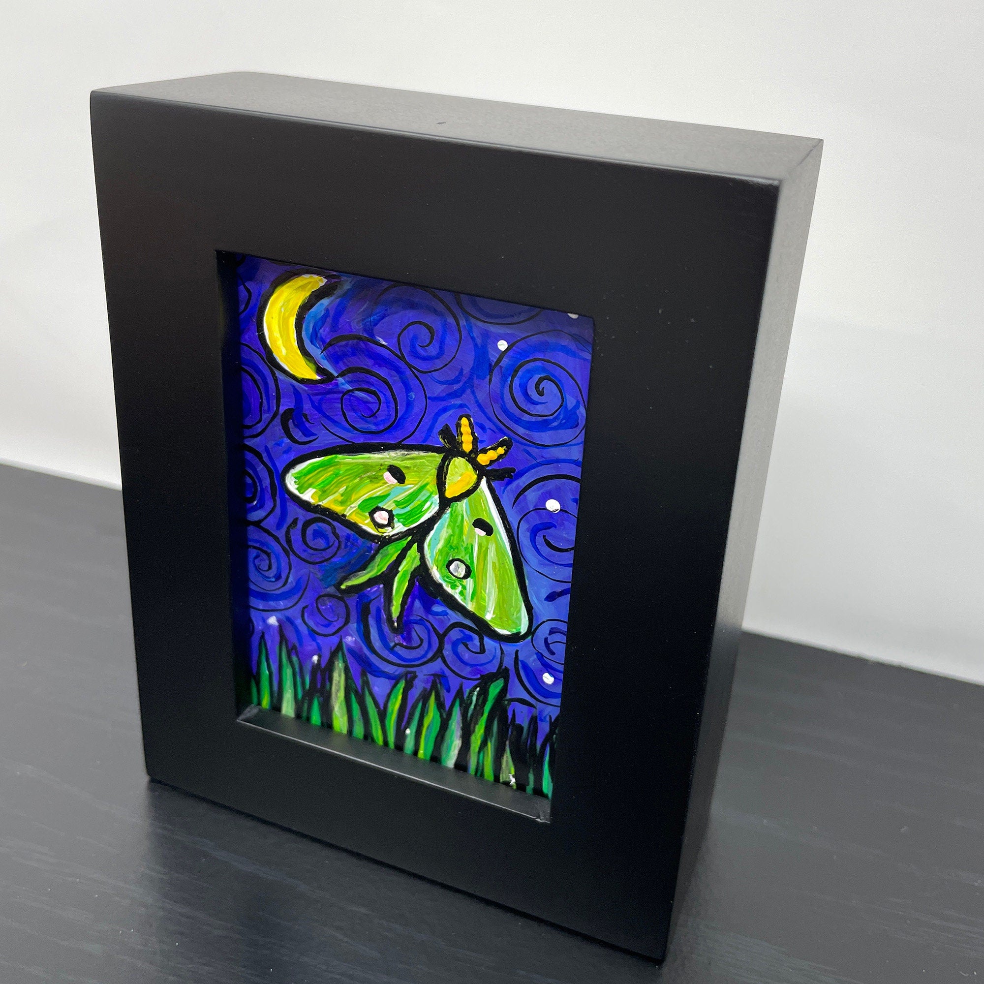 Side view of luna Moth painting in black wood frame siting on black table. Painting features green and yellow luna moth on night sky with crescent yellow moon and white stars. Long grasses dance along the bottom of the scene.