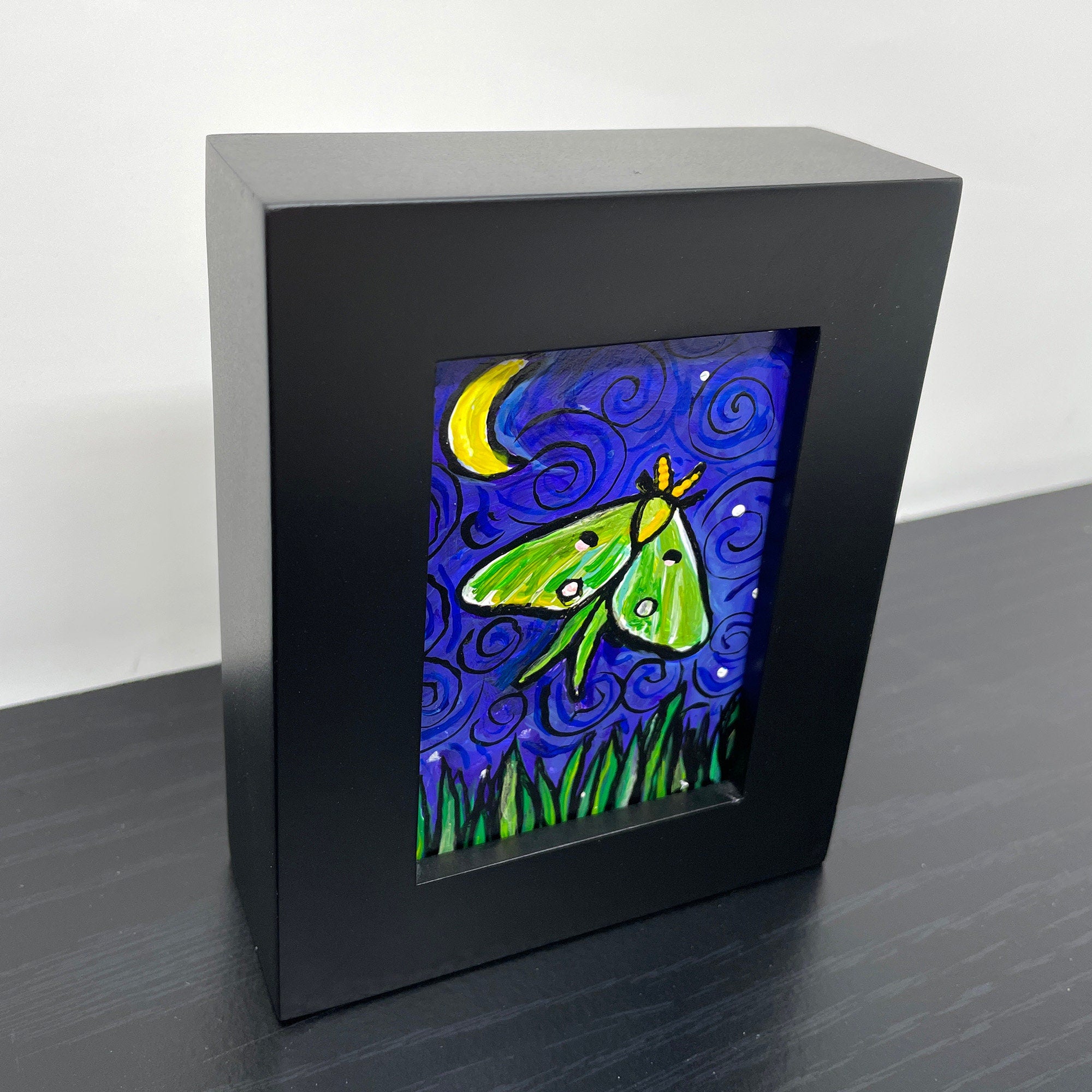 Side view of Luna Moth painting on a shelf. Luna Moth painting in black wood frame features green and yellow luna moth on night sky with crescent yellow moon and white stars. Long grasses dance along the bottom of the scene.
