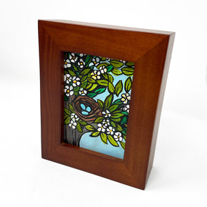 Side view of Robin&#39;s Nest painting in brown wood frame. Painting shows blue robin&#39;s eggs in a brown nest in a tree with flowers and leaves. Light blue sky is visible between the branches.