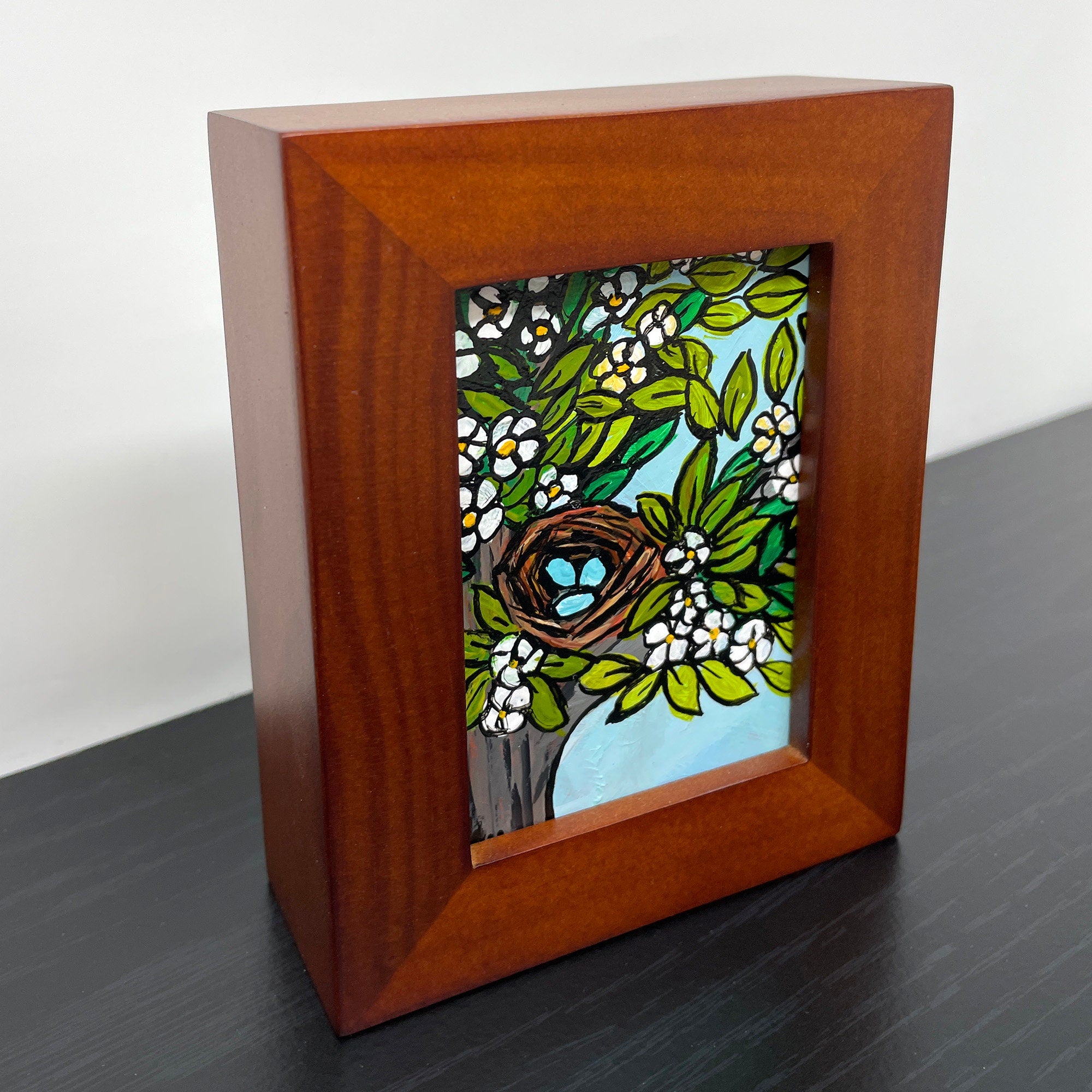 side view of Robin&#39;s Nest painting in brown wood frame. Painting shows blue robin&#39;s eggs in a brown nest in a tree with flowers and leaves. Light blue sky is visible between the branches.