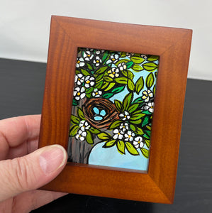 Hand holding Robin&#39;s Nest painting in brown wood frame. Painting shows blue robin&#39;s eggs in a brown nest in a tree with flowers and leaves. Light blue sky is visible between the branches.