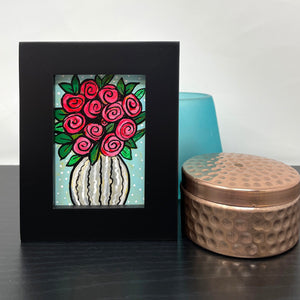 Small vase of roses painting with copper box and blue candle holder on black table. Painting features white striped vase and red roses on light blue background with white dots.