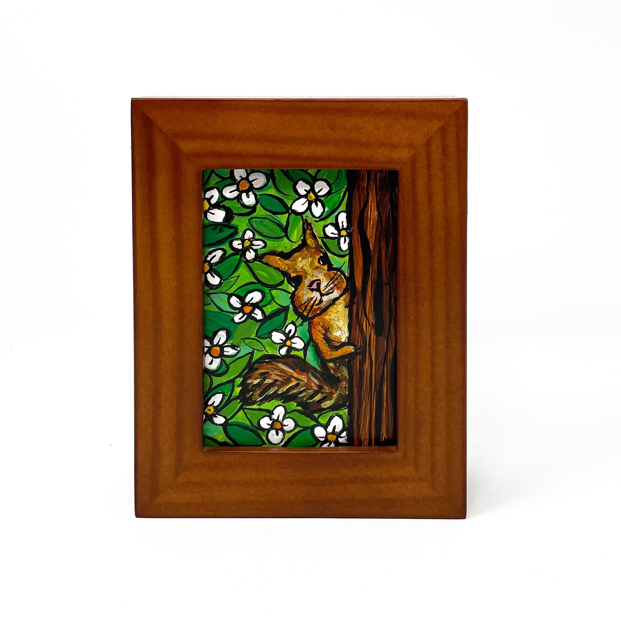 Mini squirrel painting in wood frame on white background. Painting shows squirrel peeking behind a tree at the viewer. Background is white flowers and green leaves.