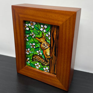 Side view of mini squirrel painting in wood frame on black table. Painting shows squirrel peeking behind a tree at the viewer. Background is white flowers and green leaves.