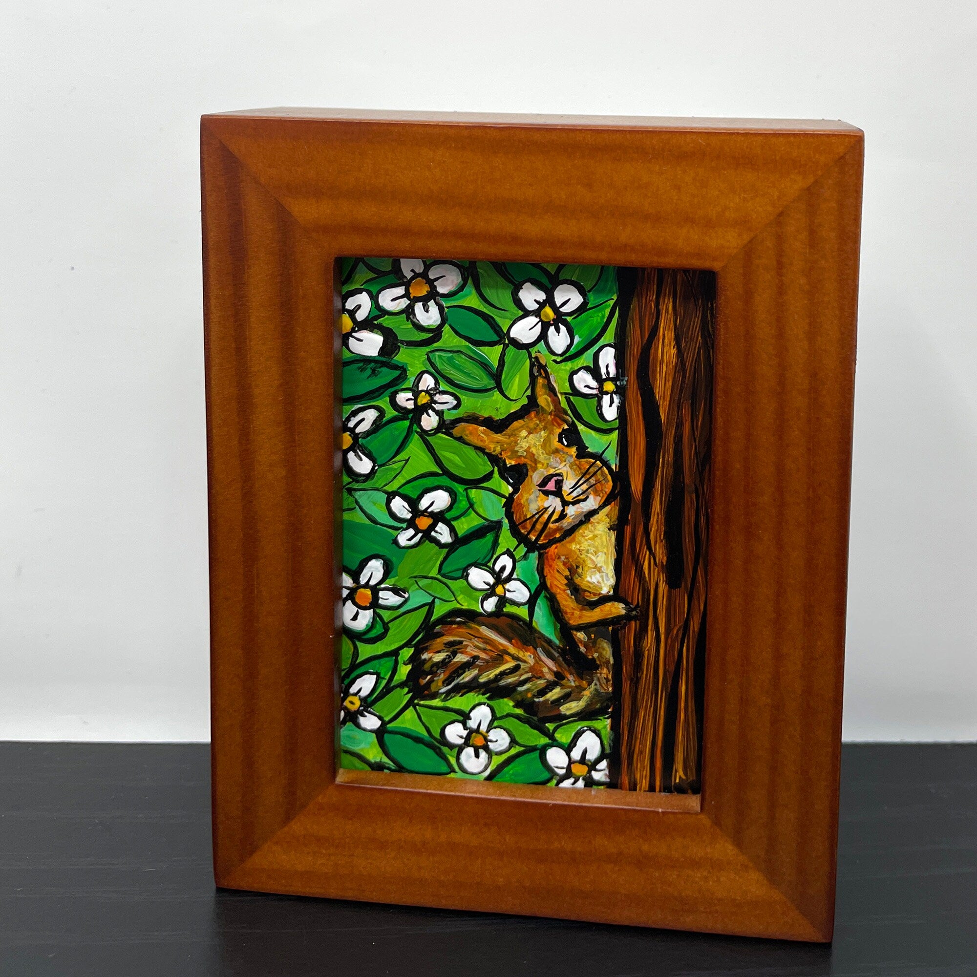 Front view of mini squirrel painting in wood frame on black table. Painting shows squirrel peeking behind a tree at the viewer. Background is white flowers and green leaves.