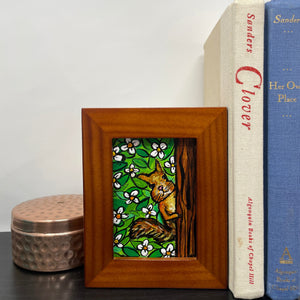 Mini squirrel painting in wood frame on shelf with books and trinket box. Painting shows squirrel peeking behind a tree at the viewer. Background is white flowers and green leaves.
