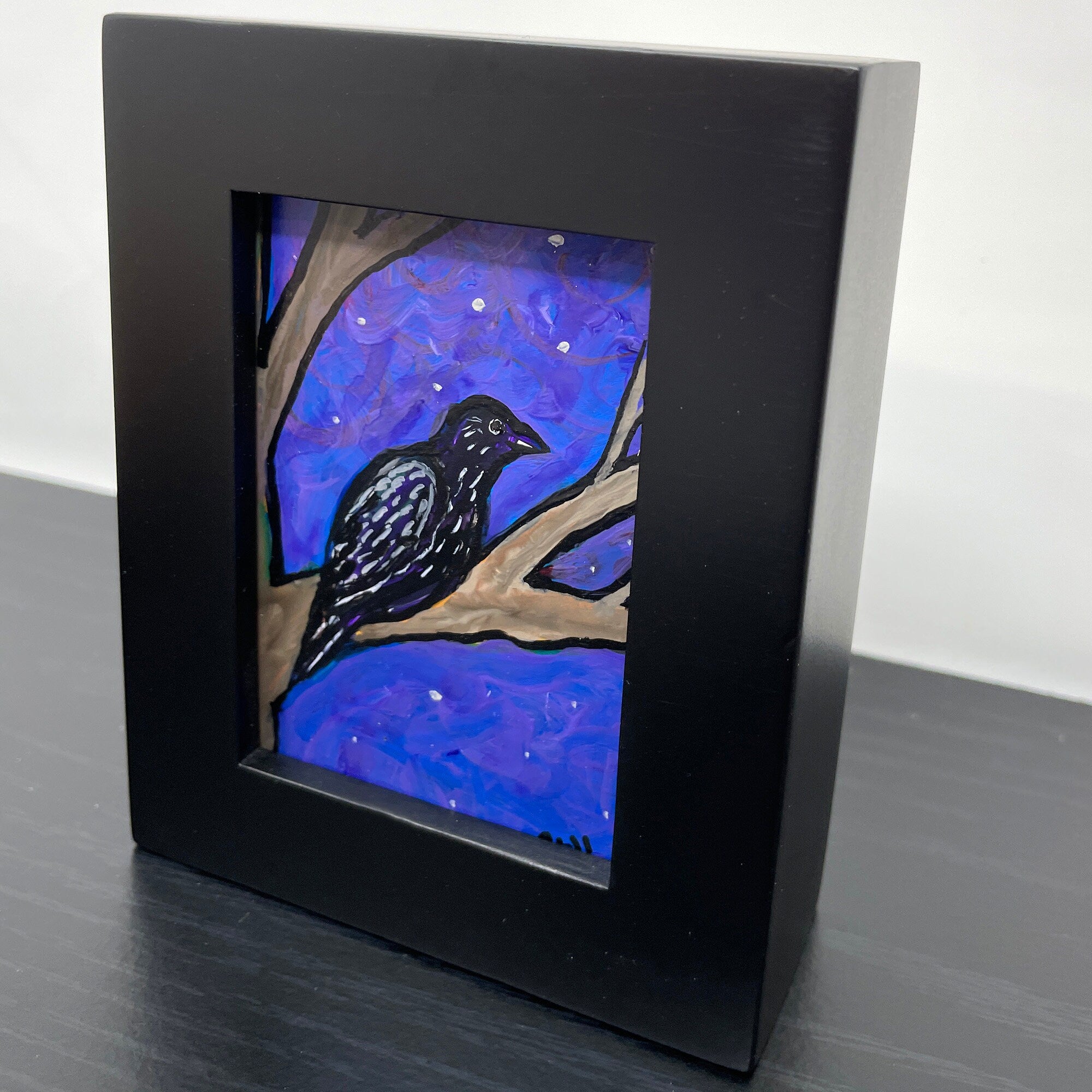 Side view of Raven painting in black frame. Raven is sitting on tree with night sky behind him.