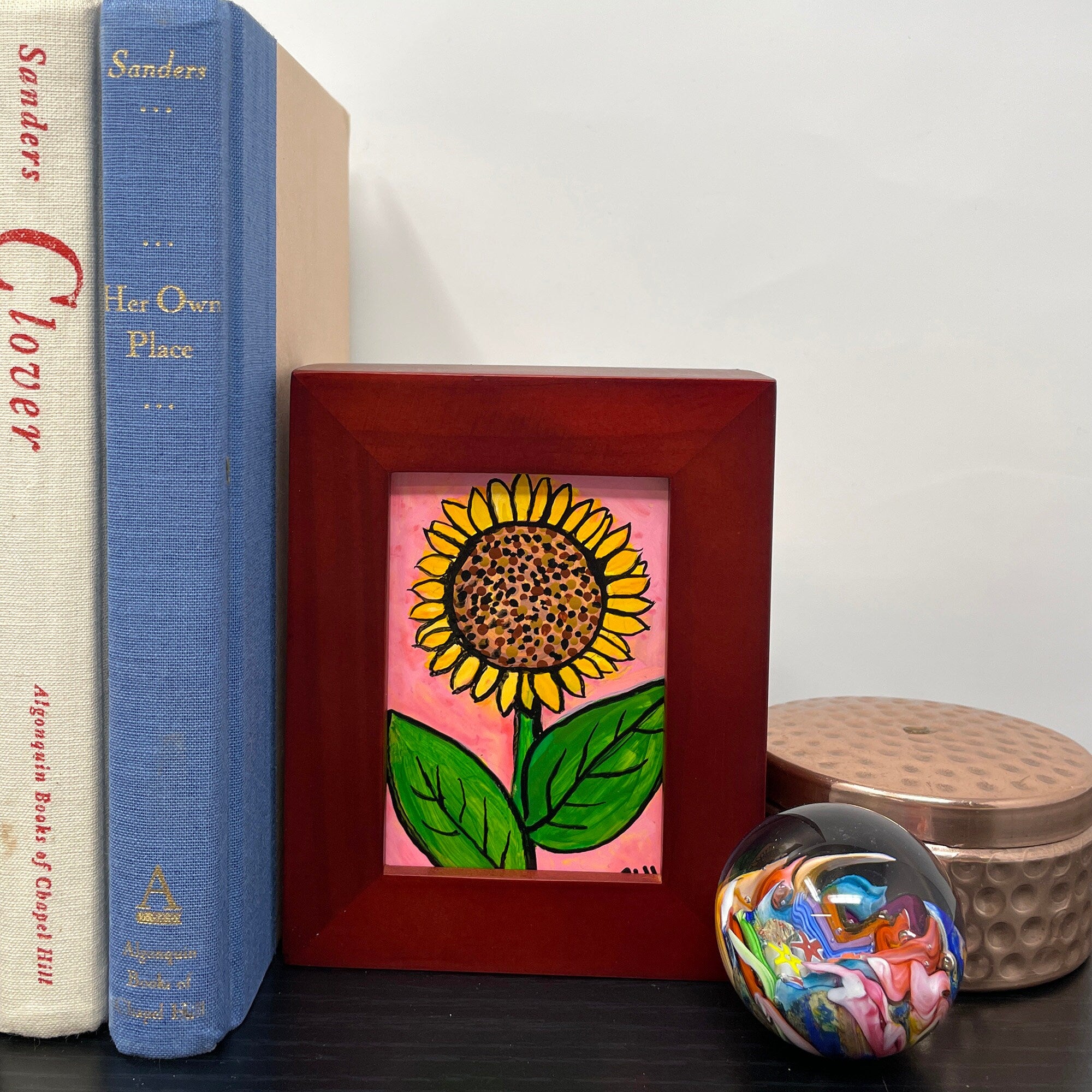 Bookshelf featuring small sunflower painting in cherry wood frame. Sunflower with brown center, yellow petals, and green leaves on pink background