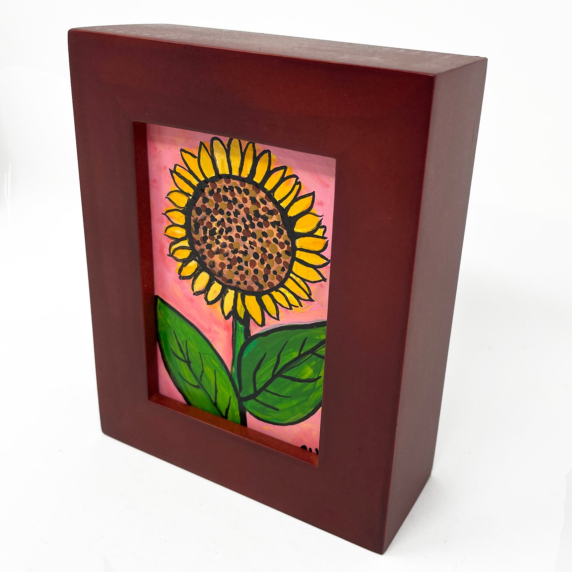 Side view of Sunflower Day painting in wood frame. Painting shows sunflower in the center with yellow petals, brown center, and green leaves on light pink background.