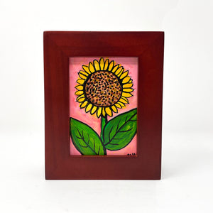 Front view of Sunflower Day painting in wood frame. Painting shows sunflower - yellow petals, brown center with dots, and green leaves on light pink.