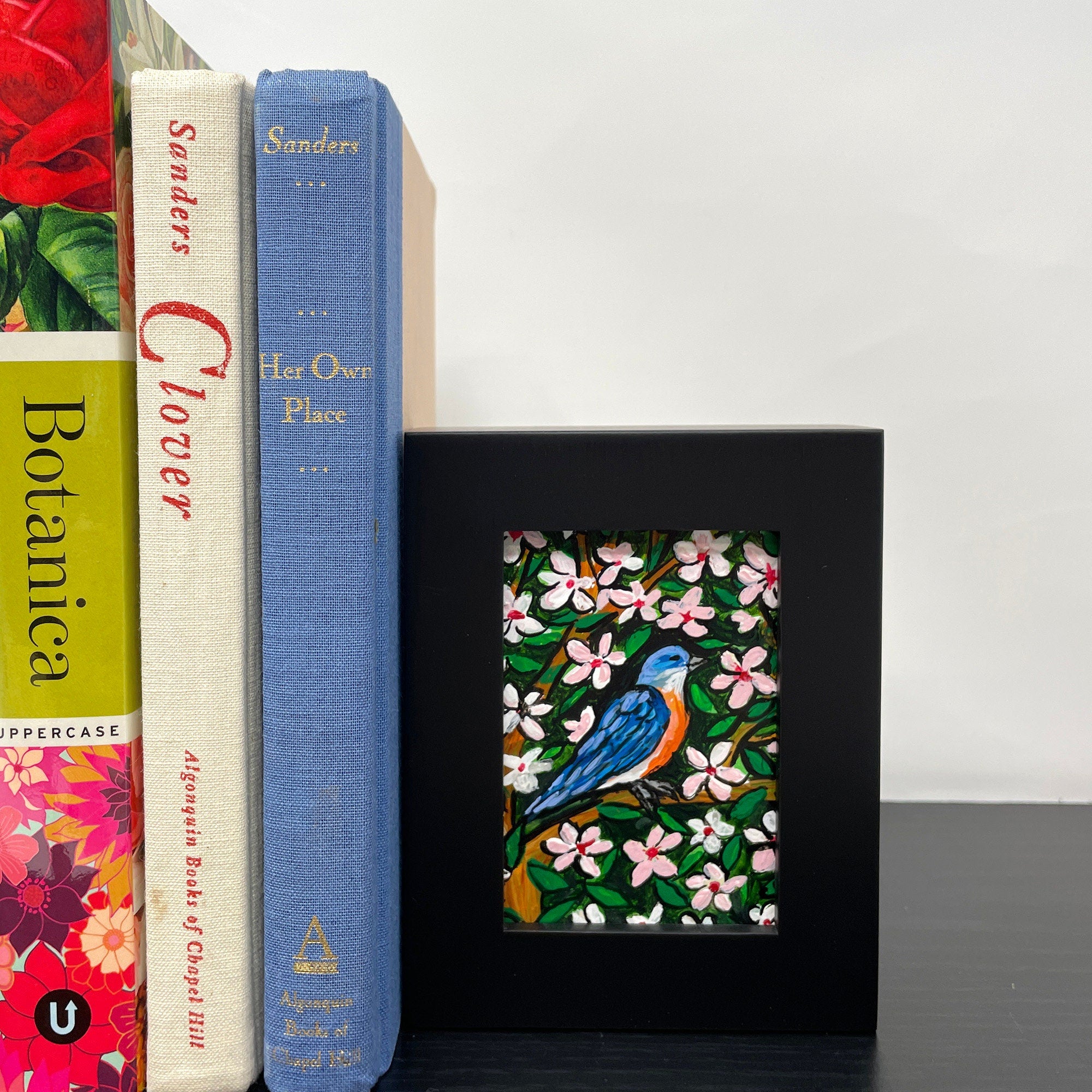 Framed bluebird painting sitting on a black shelf with books. In the painting a bluebird sits on a cherry tree surrounded by pink and white cherry blossoms and green leaves.