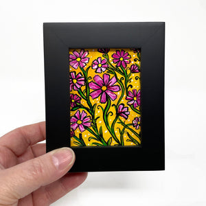 Hand holding black wood frame with pink cosmos flower painting. Flowers and green leaves are on a yellow ochre background.